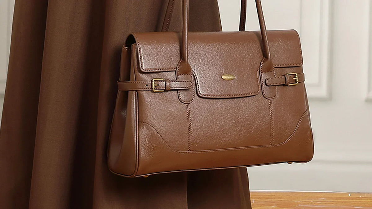 Close view of a brown leather handbag against a brown skirt, showing its flap, buckles, and stitching.
