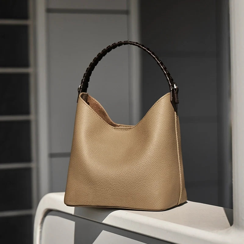 Pebbled leather bucket bag with a black braided handle on a white windowsill