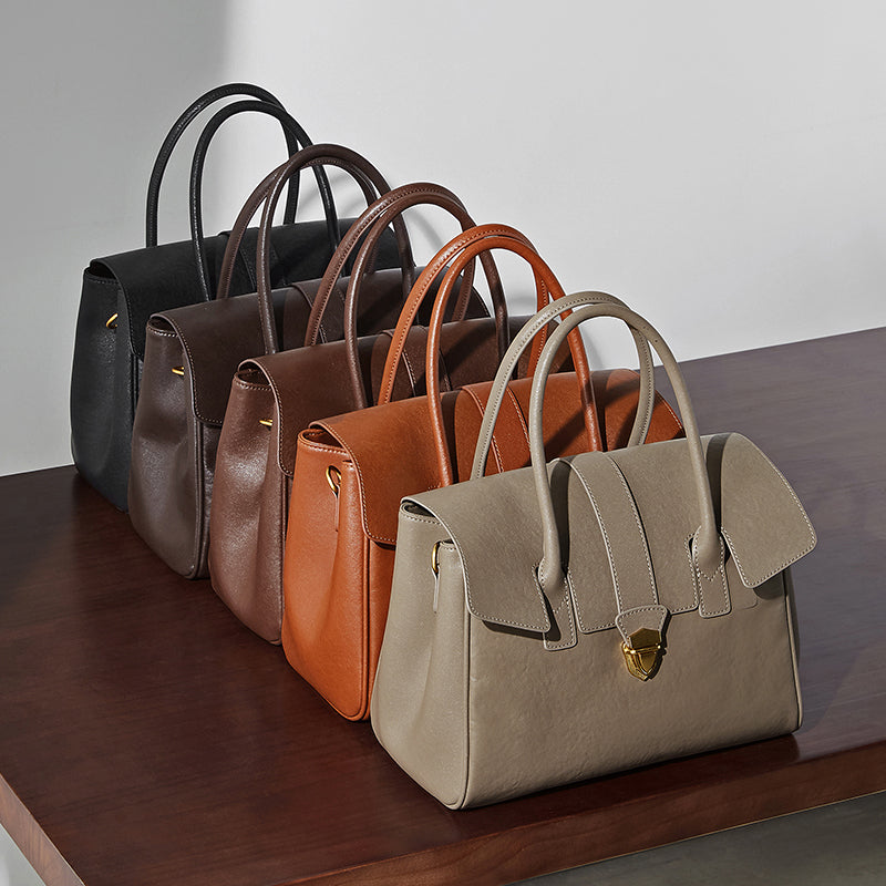 Four leather top-handle satchels in black, dark brown, brown, and gray on a wooden table