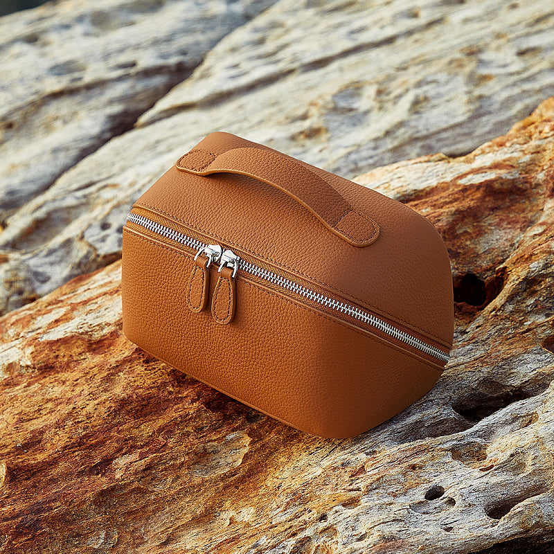 Brown pebbled leather makeup bag with a top handle and double zipper resting on a rock