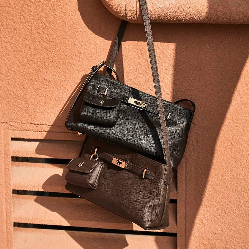 Two Cuirae genuine leather crossbody bags in black and brown with small attached pouches hanging against a textured terracotta wall in the sunlight.