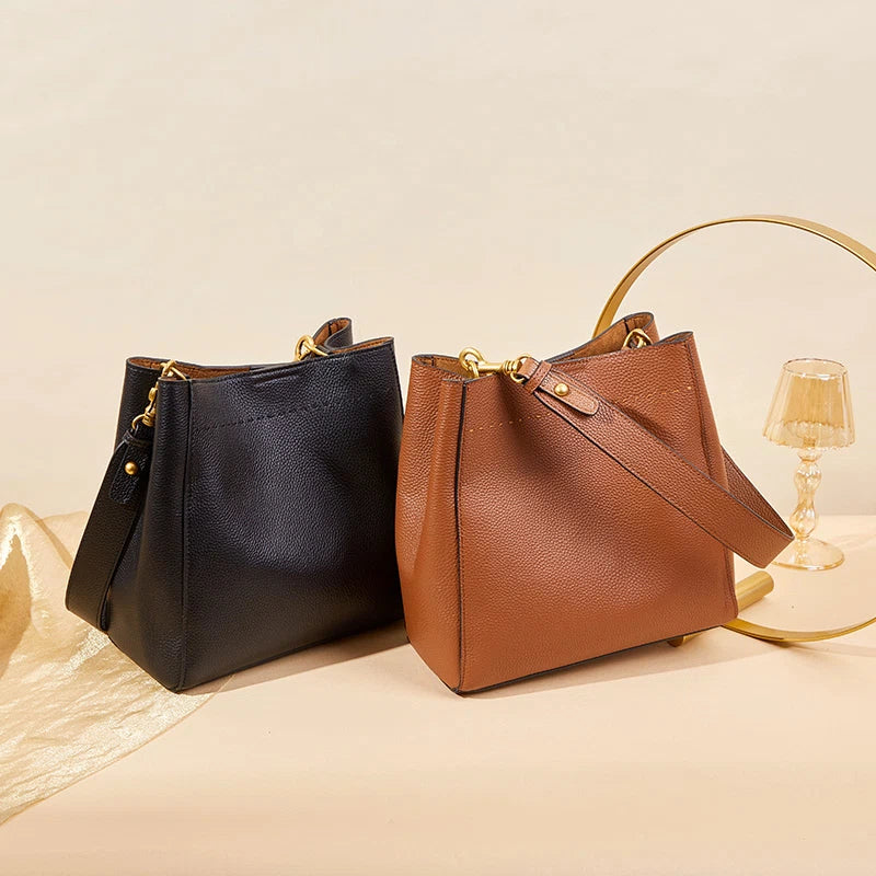 Two Cuirae top grain cowhide bucket bags in black and brown colors resting on a light surface next to a gold decorative ring.