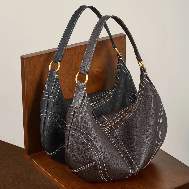 Two Cuirae top grain cowhide shoulder bags in black and dark brown with white stitching sitting on a wooden stand.
