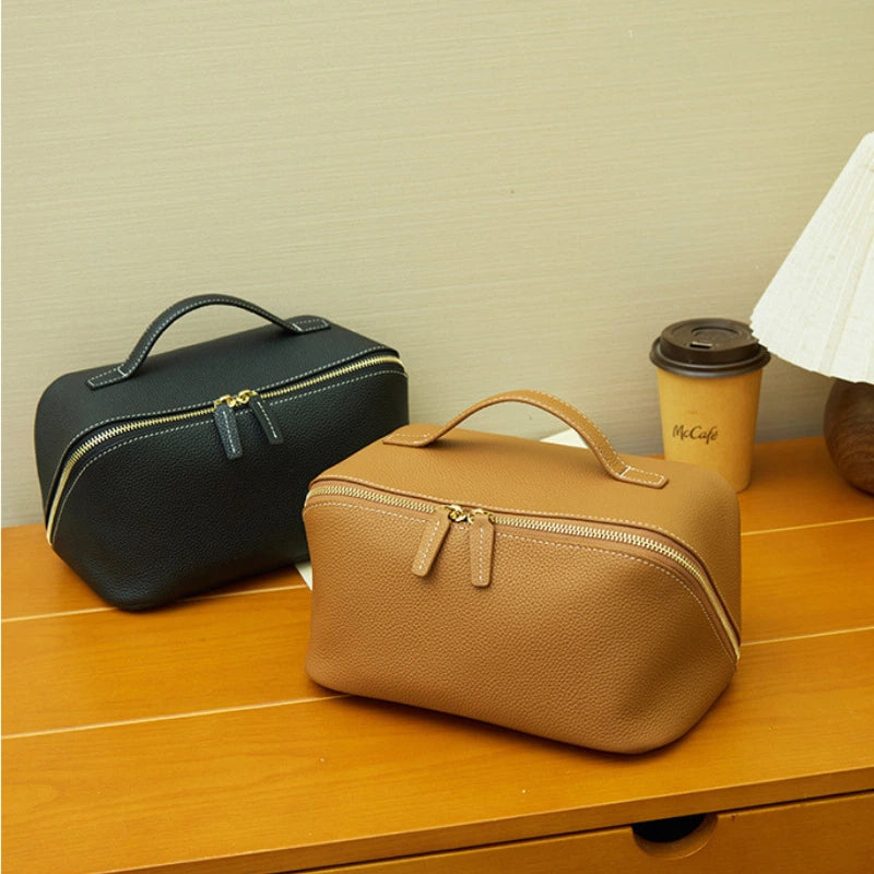 Two Cuirae top grain cowhide makeup bags in black and brown resting on a wooden desk next to a coffee cup and a lamp.