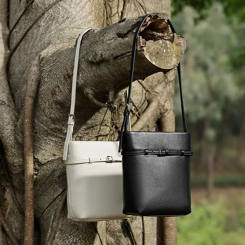 Two Cuirae top grain cowhide bucket bags in black and white hanging from a tree branch outside.