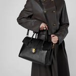Model holding Cuirae black genuine leather satchel bag with top handles, showing adjustable crossbody strap