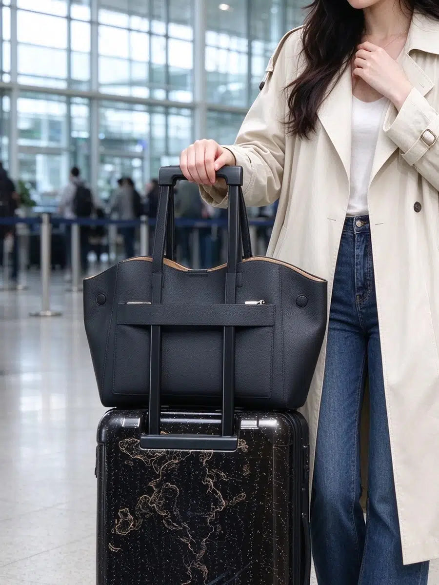 Cuirae black genuine leather tote bag secured to a rolling suitcase handle with its rear luggage sleeve at an airport