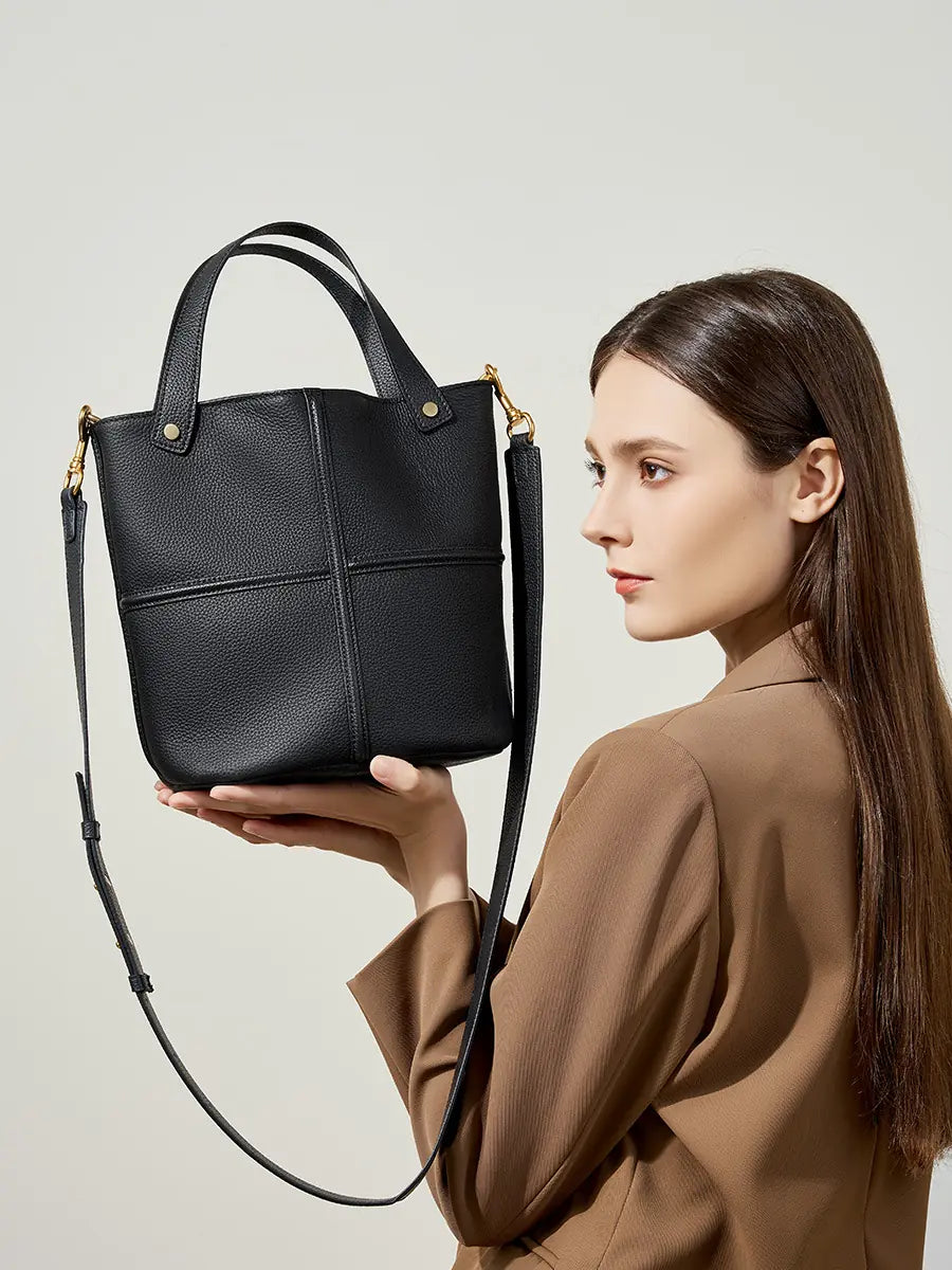 Model holding up a black leather bucket bag, showing its raised cross seams and long shoulder strap
