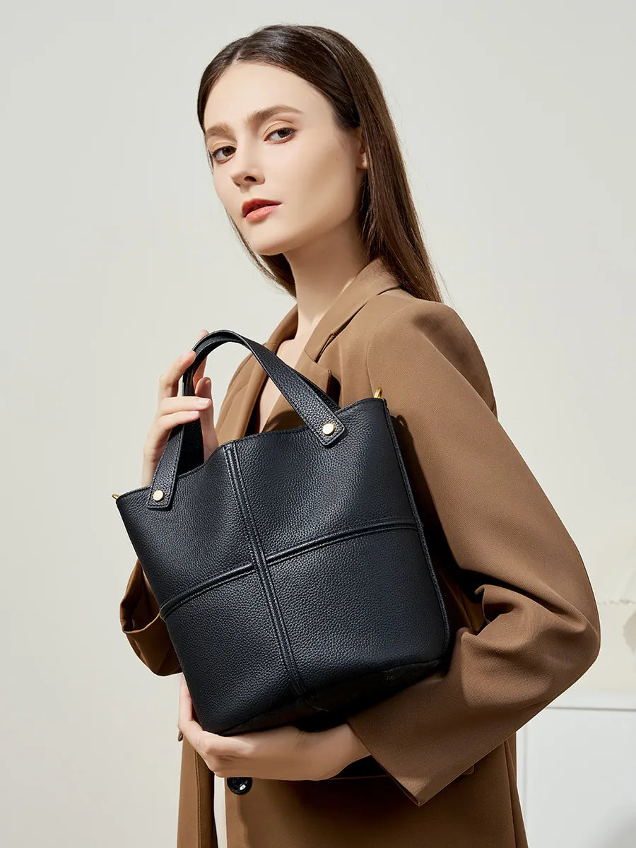 Model in a brown blazer holding a black pebbled leather bucket bag with twin handles and gold-tone rivets