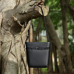 Cuirae black pebbled leather bucket bag hanging on tree branch, outdoor nature scene showing belt buckle detail