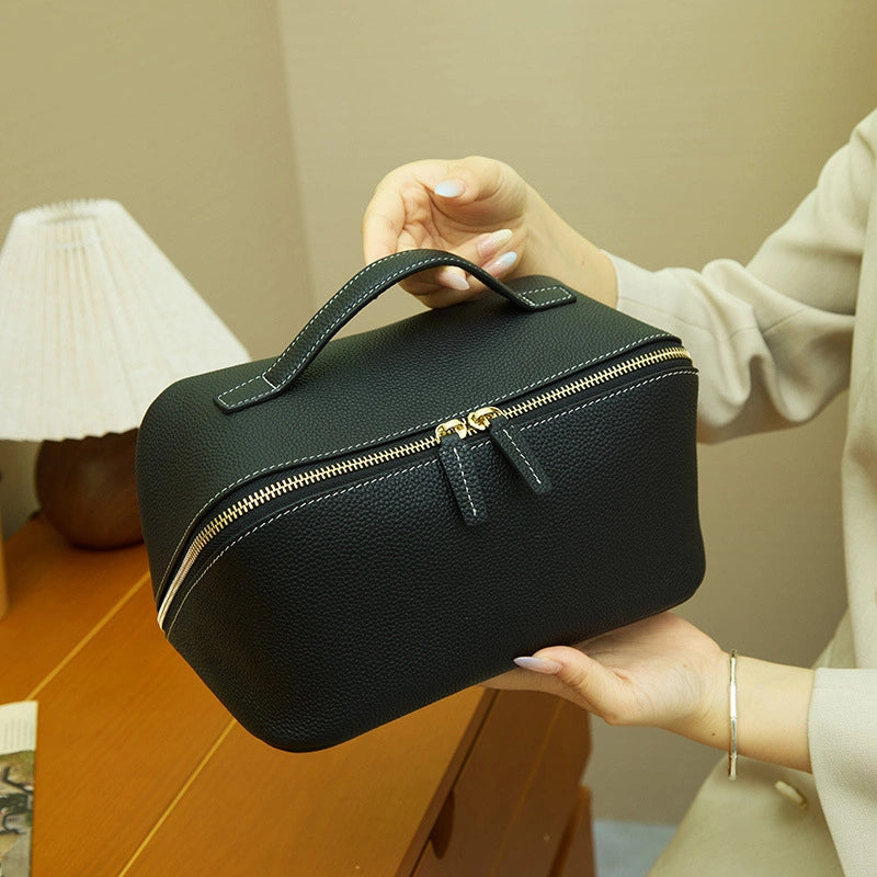 Cuirae black pebbled genuine leather makeup bag with top handle, hand held by model showing gold zipper details