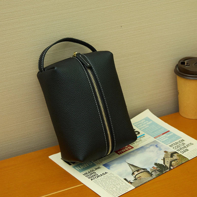 A Cuirae black pebbled leather toiletry bag with a vertical zipper and top handle resting on a newspaper on a wooden desk.