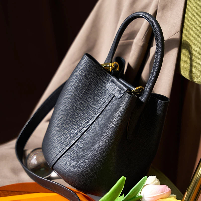 A Cuirae black top grain cowhide bucket bag with a shoulder strap and top handle resting on a chair next to tulips.