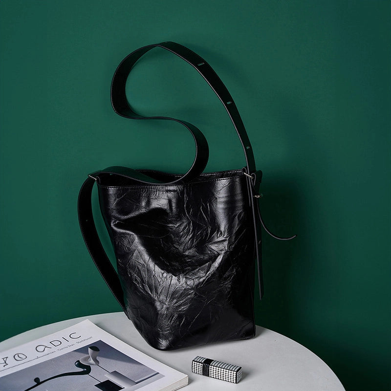 A Cuirae black top grain cowhide bucket bag with a long shoulder strap resting on a white table against a dark green wall.