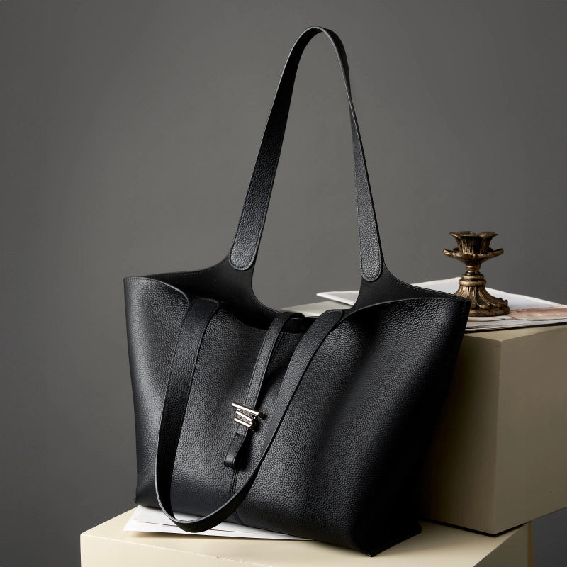 A Cuirae black top grain cowhide tote bag with long shoulder straps resting on a pedestal next to a decorative metal piece against a grey background.