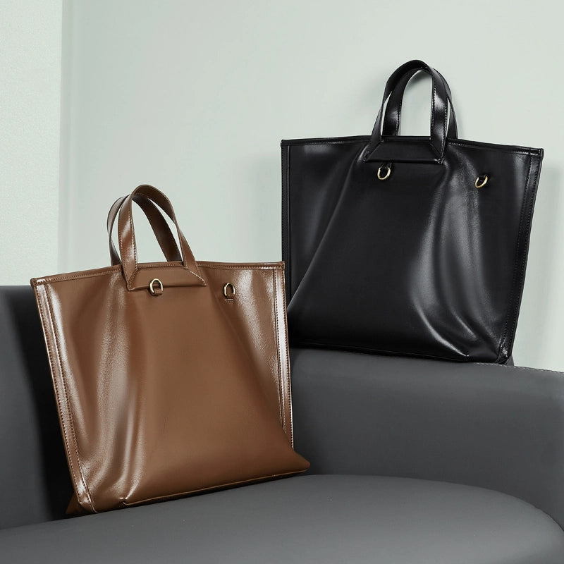 Two Cuirae patent leather tote bags in brown and black resting on a grey sofa.