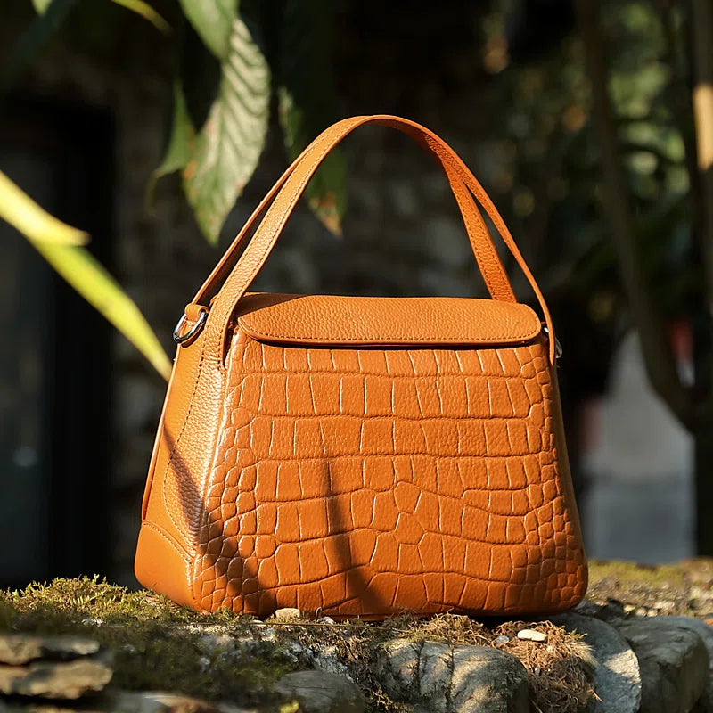 A Cuirae brown crocodile-pattern genuine leather crossbody bag with a top handle resting on a mossy stone wall.