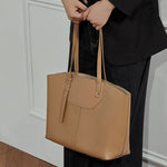 Model stands against the wall with her hands by her sides, holding a Cuirae brown genuine leather bag.