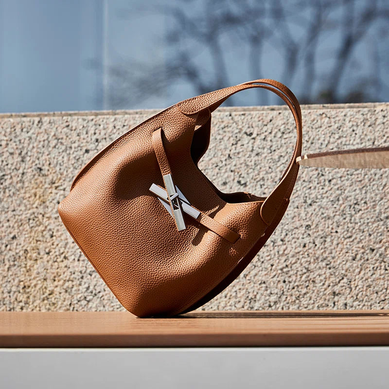 Cuirae brown genuine leather crossbody bag on outdoor stone bench, angled view showing pebbled texture and cross turnlock
