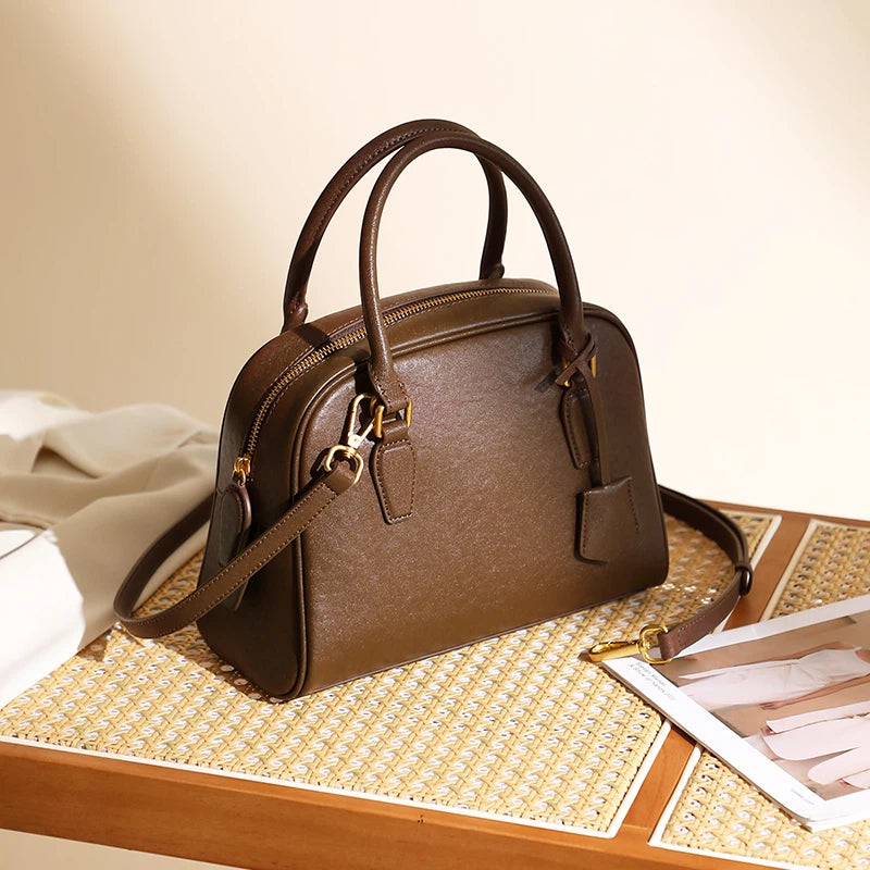 A Cuirae brown genuine leather handbag with a shoulder strap resting on a rattan table next to a magazine.