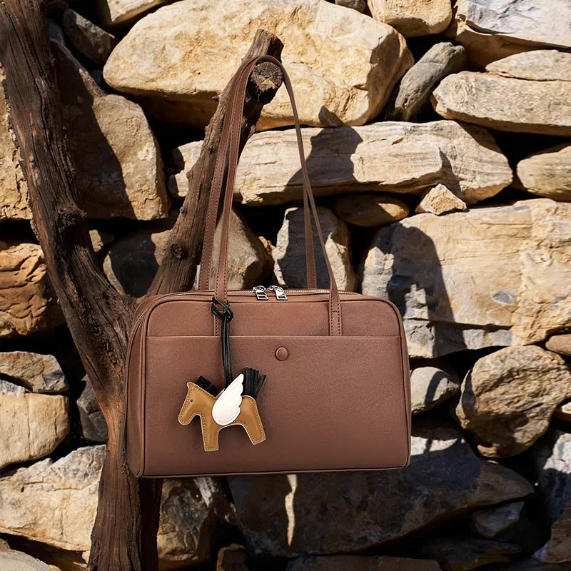 A Cuirae brown genuine leather shoulder bag with a horse charm hanging on a dry tree branch against a stone wall.