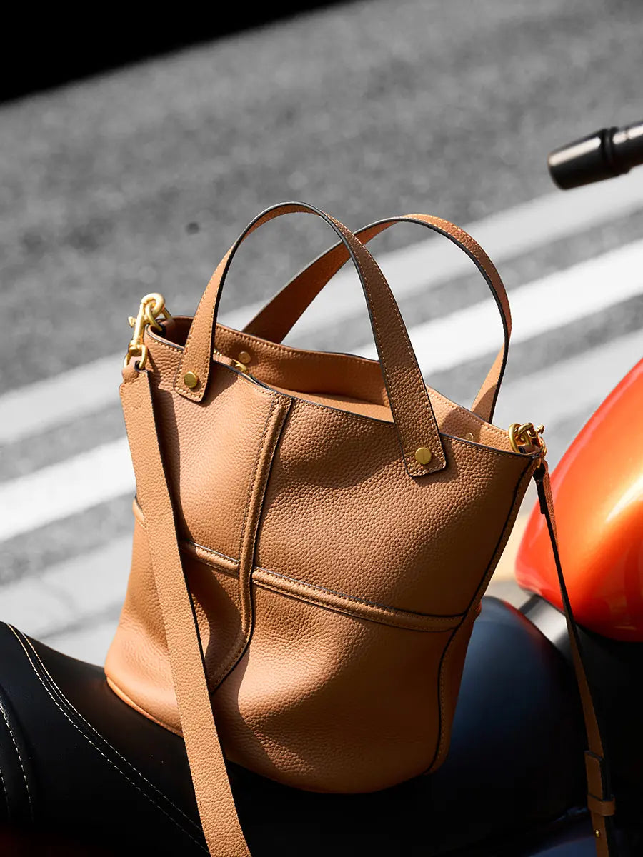 Angled view of a brown leather bucket bag on a motorcycle seat, showing its pebbled texture and rounded base