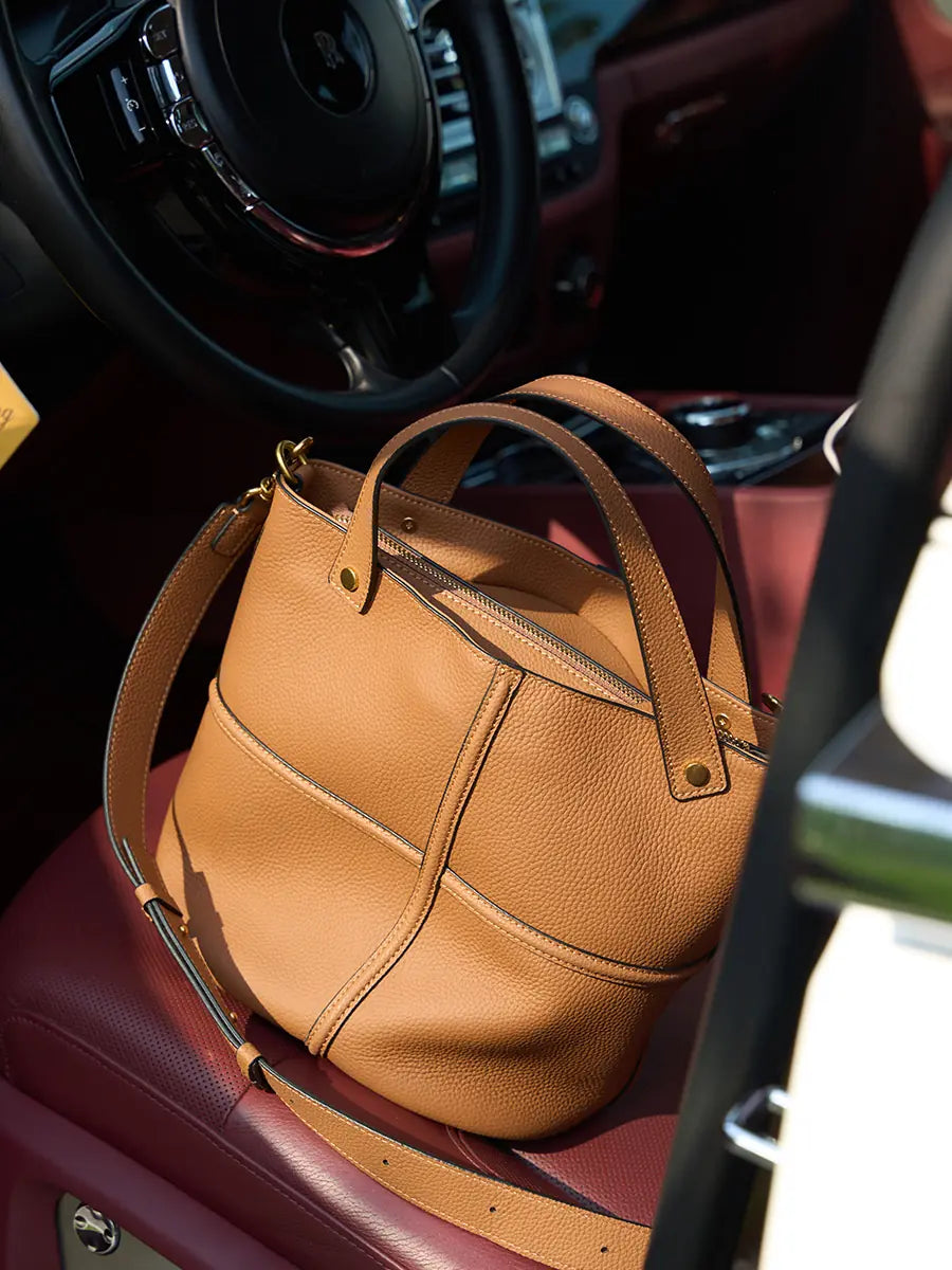 Brown leather bucket bag on a car seat, showing its zip closure, cross seams, and adjustable strap