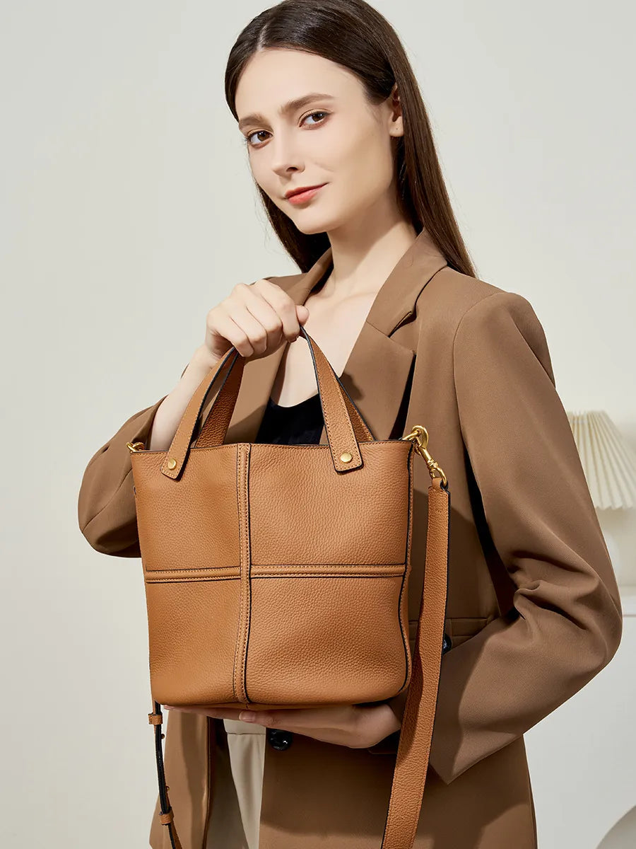 Model holding a brown pebbled leather bucket bag in front of her, showing its rounded shape and cross-seam detailing