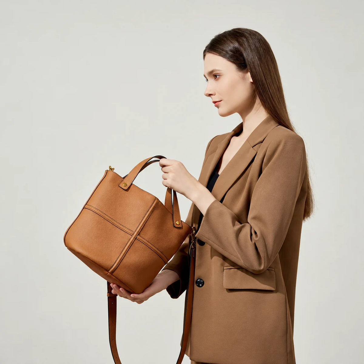 Side view of a model holding a brown pebbled leather bucket bag with raised cross seams and a detachable shoulder strap.