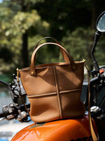 Brown leather bucket bag displayed on an orange motorcycle, with raised cross seams and gold-tone hardware