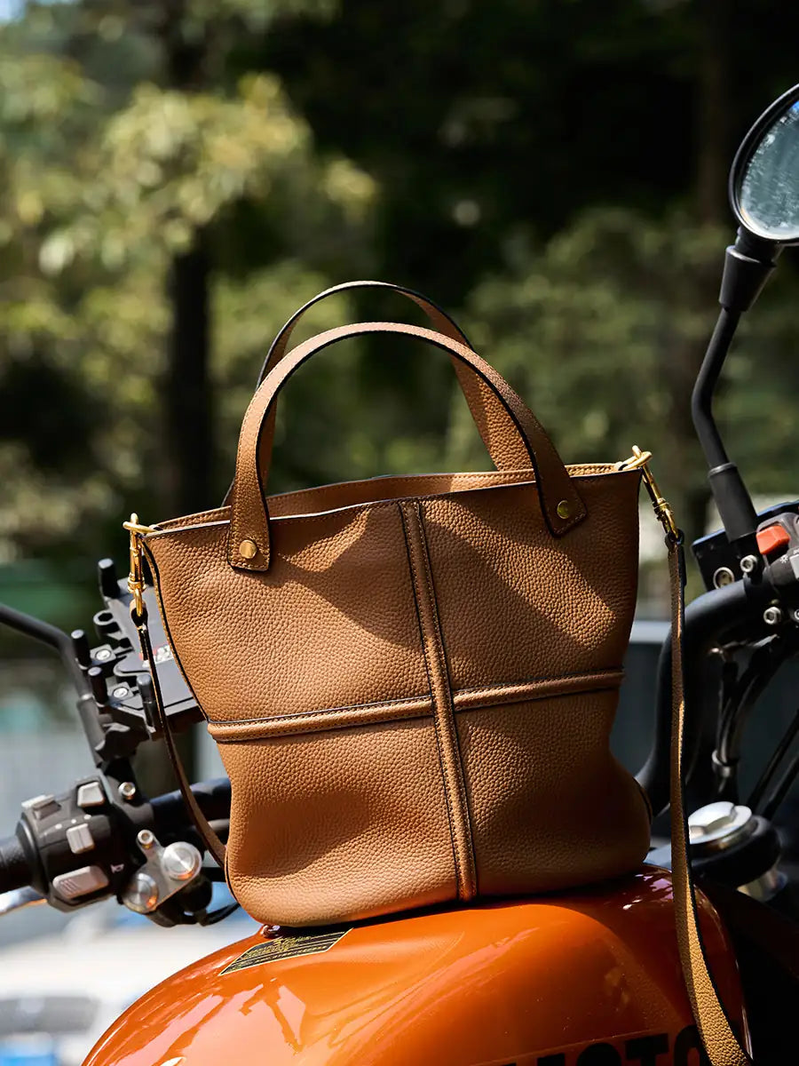 Brown leather bucket bag displayed on an orange motorcycle, with raised cross seams and gold-tone hardware