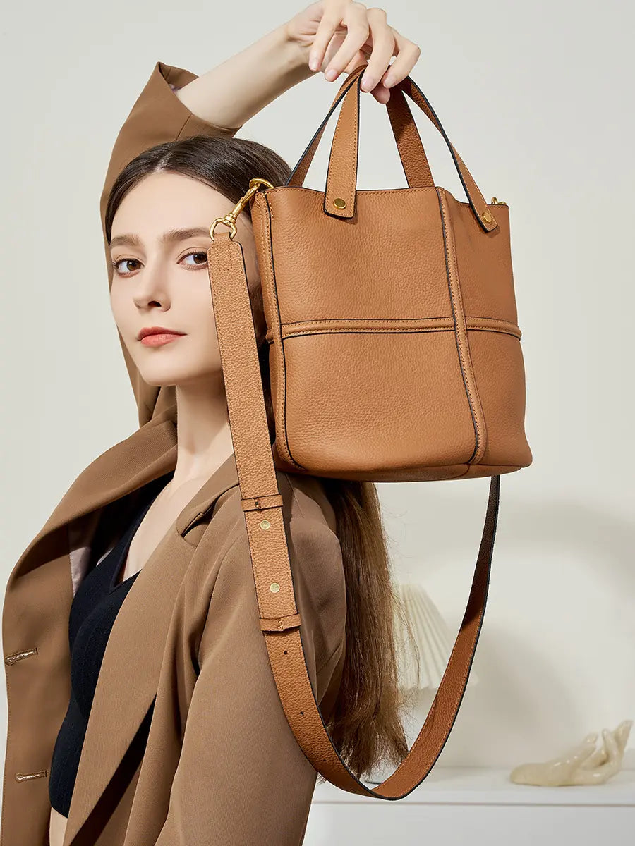 Model lifting a brown leather bucket bag by its twin handles, with the adjustable shoulder strap hanging below