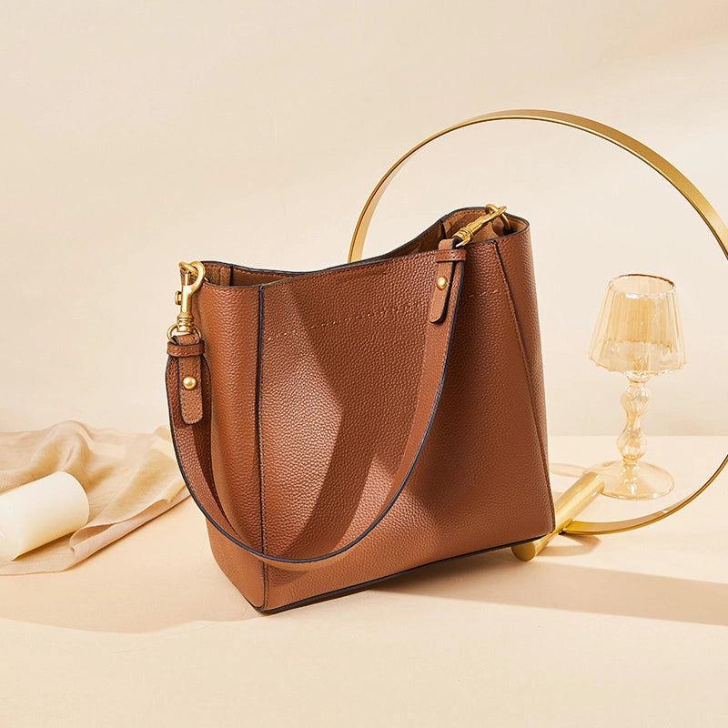 Cuirae brown pebbled leather bucket bag on warm beige table, still life with gold decor and glass goblet