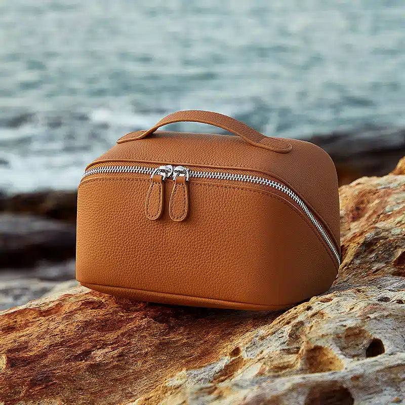 A Cuirae brown pebbled leather makeup bag with a top handle and double zippers resting on a textured rock with the sea in the background.