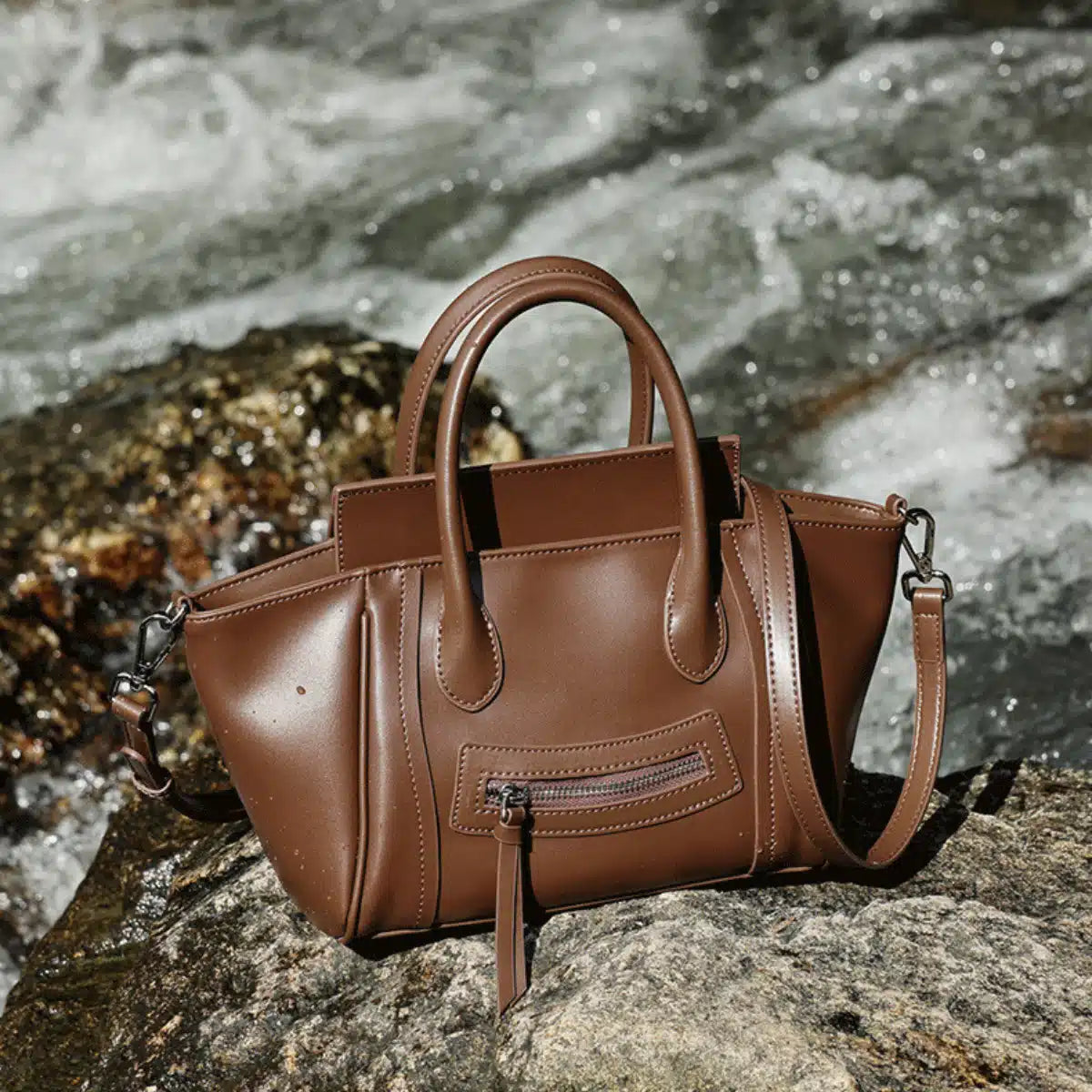 A Cuirae brown top grain cowhide handbag resting on a rock with rushing river water in the background.
