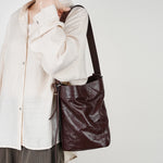 Woman wearing Cuirae burgundy crinkled leather bucket bag on shoulder, hand resting on strap with beige linen shirt outfit