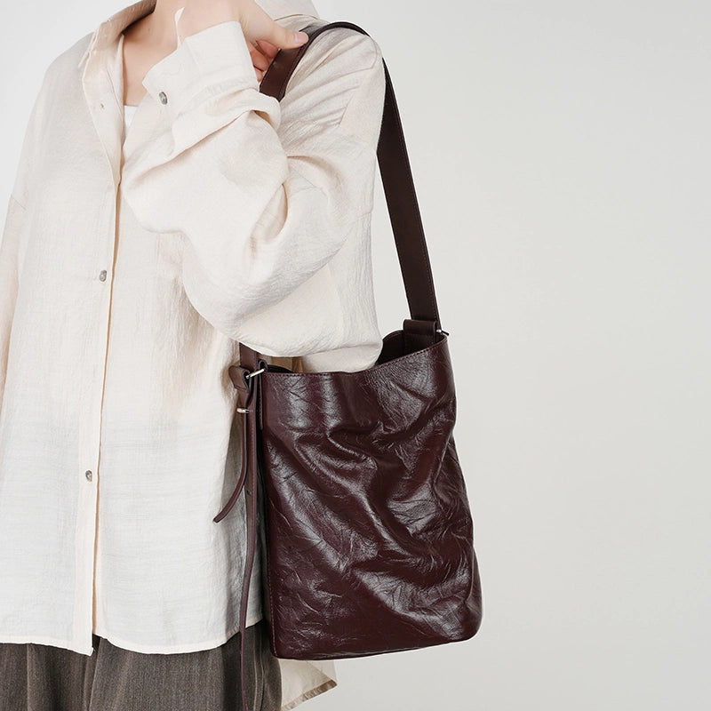 Woman wearing Cuirae burgundy crinkled leather bucket bag on shoulder, hand resting on strap with beige linen shirt outfit