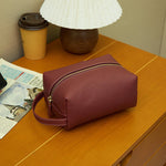 Cuirae burgundy pebbled leather makeup bag on wooden nightstand, showing gold zipper and wrist strap