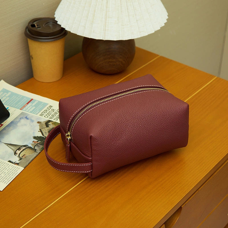 Cuirae burgundy pebbled leather makeup bag on wooden nightstand, showing gold zipper and wrist strap