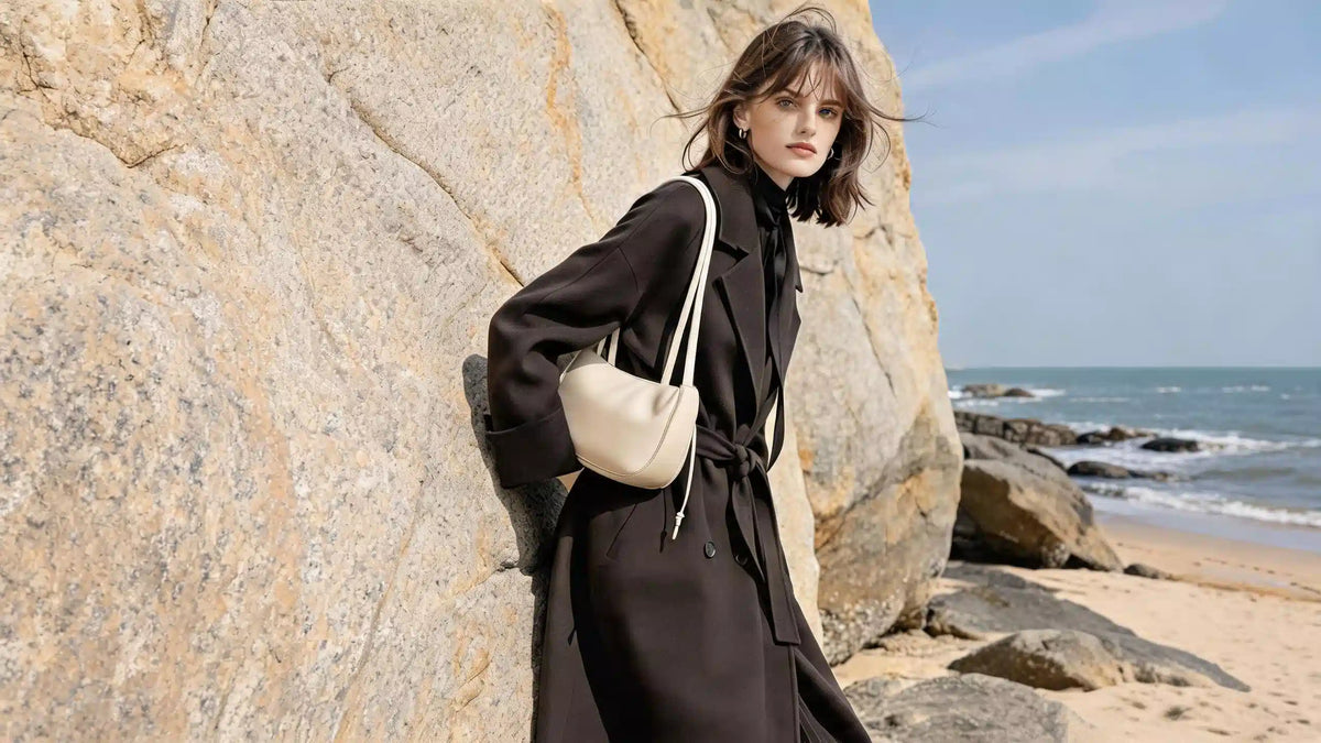 Woman wearing a cream Cuirae crescent shoulder bag beside coastal rocks.
