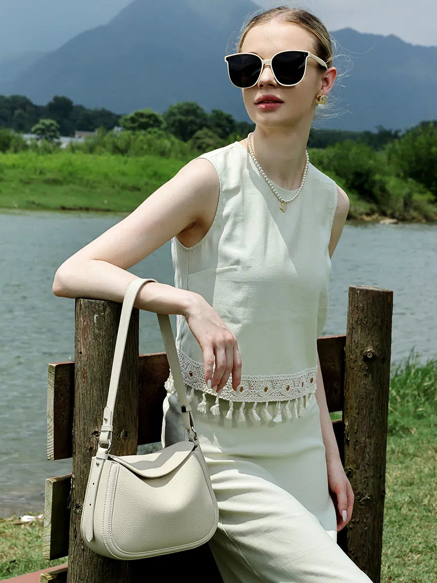 Model carrying a Cuirae ivory pebbled leather shoulder bag on her forearm beside a wooden fence.