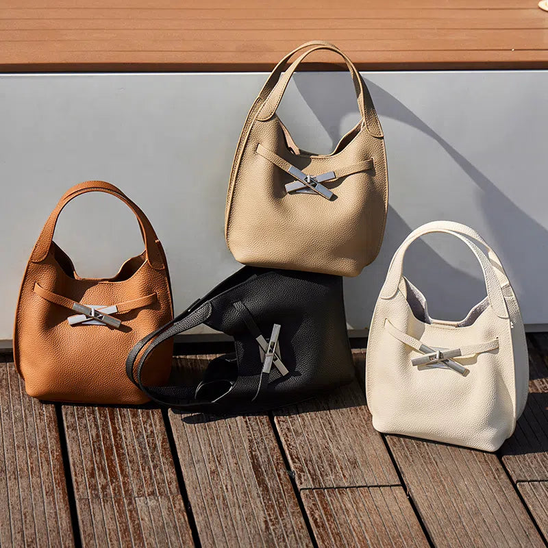 Four Cuirae genuine leather crossbody bags in tan, beige, black, and cream with silver clasps resting on a wooden deck in the sunlight.