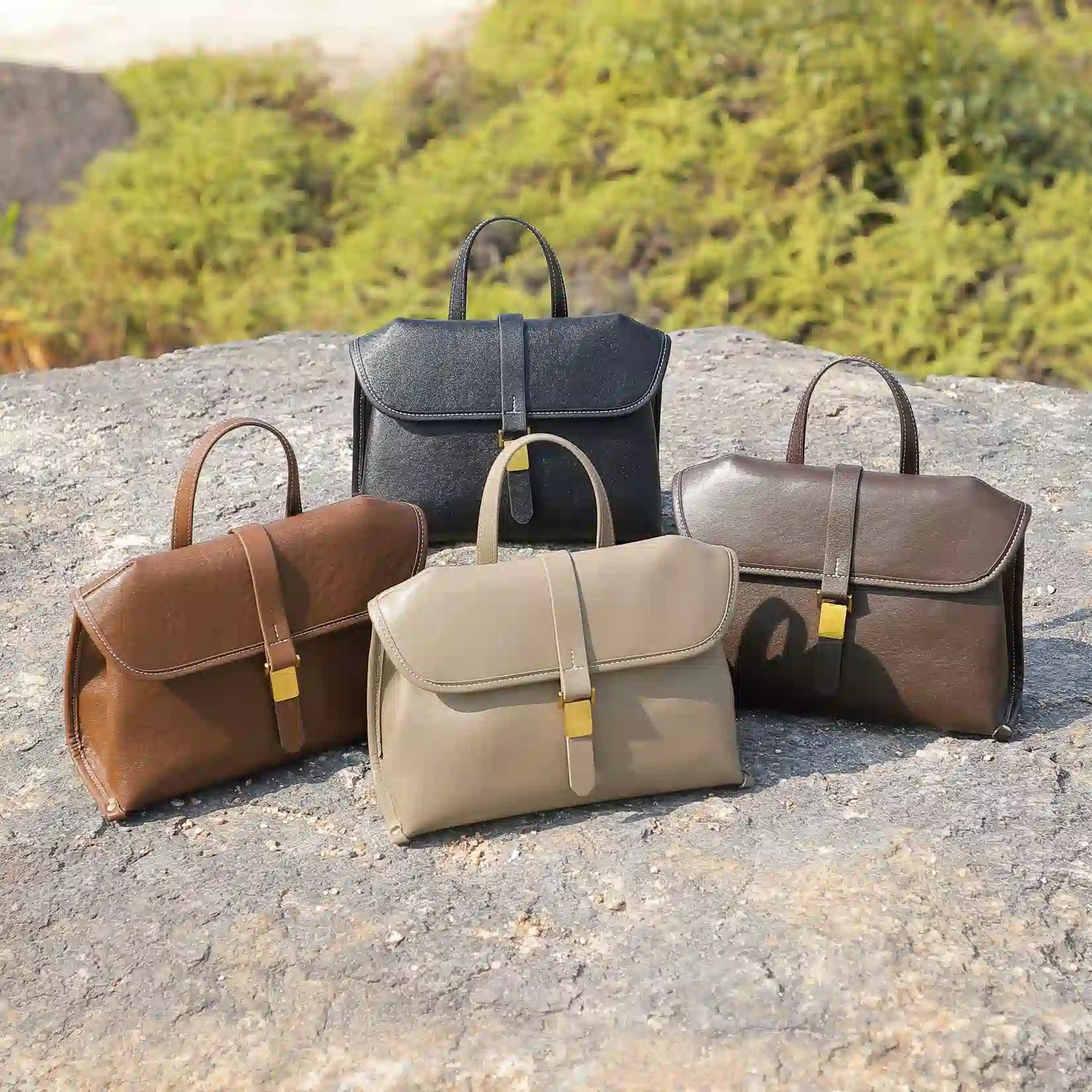 Four Cuirae genuine leather handbags in different colors resting on a large rock outside.