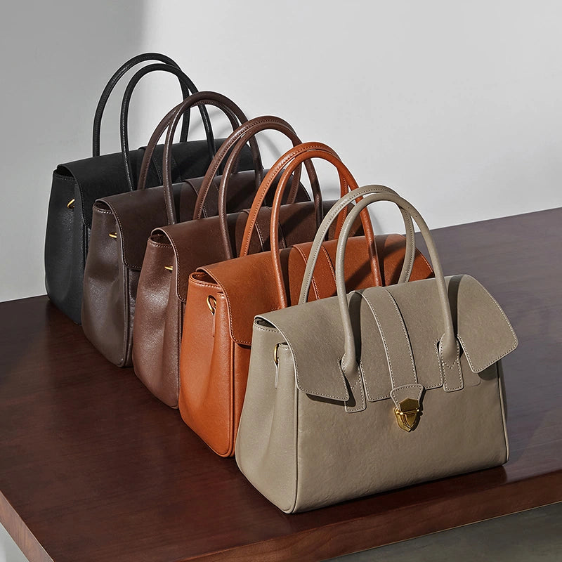 Five Cuirae genuine leather tote bags in black, dark brown, brown, tan, and taupe lined up in a row on a dark wooden table.