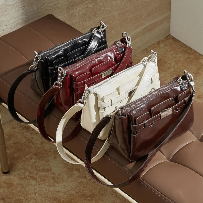 Four Cuirae patent leather crossbody bags in black, burgundy, white, and brown resting in a row on a bench.
