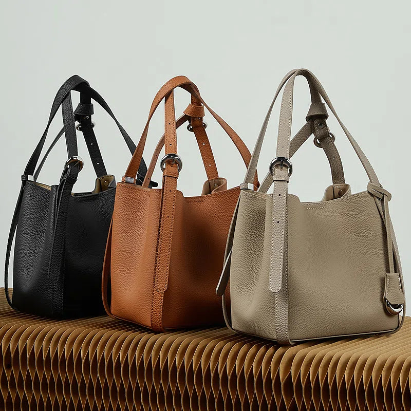 Three Cuirae top grain cowhide bucket bags in black, brown, and taupe resting on a textured bench.