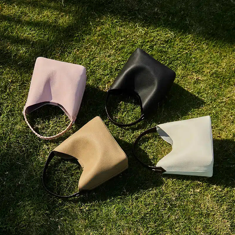 Four Cuirae top grain cowhide bucket bags in pink, black, tan, and cream resting on green grass in the sun.