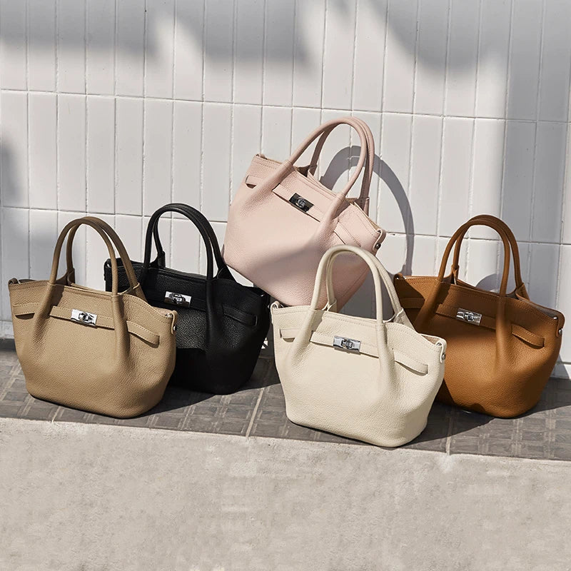 Five Cuirae top grain cowhide crossbody bags in taupe, black, pink, cream, and brown resting together against a white tiled wall.