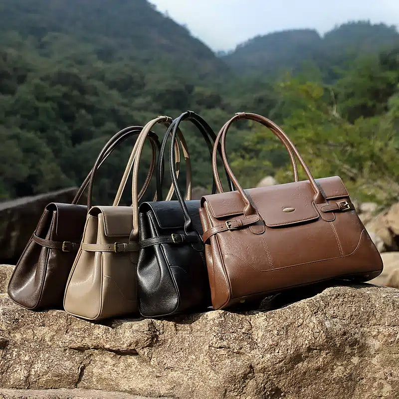 Four Cuirae top grain cowhide handbags in brown, taupe, black, and brown resting together on a large rock in a natural outdoor setting.