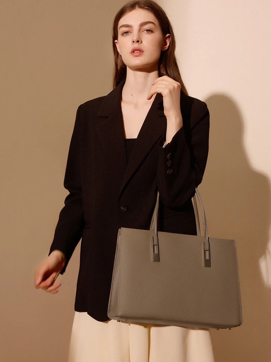 Model carrying a taupe structured leather tote on her forearm, styled with a black blazer